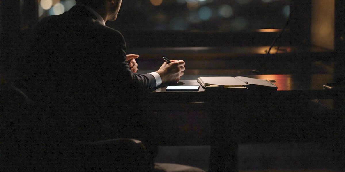 Deux personnes en entretien de part et d’autre d’un bureau sombre, un téléphone posé à plat entre elles, écran allumé.
